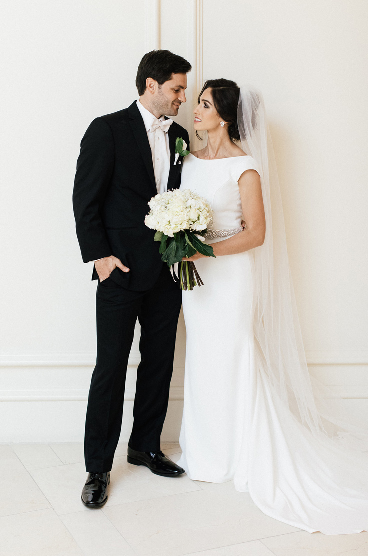 Bride in a boat neck wedding dress with a beaded belt and veil, holds a white bouquet and smiles at groom.
