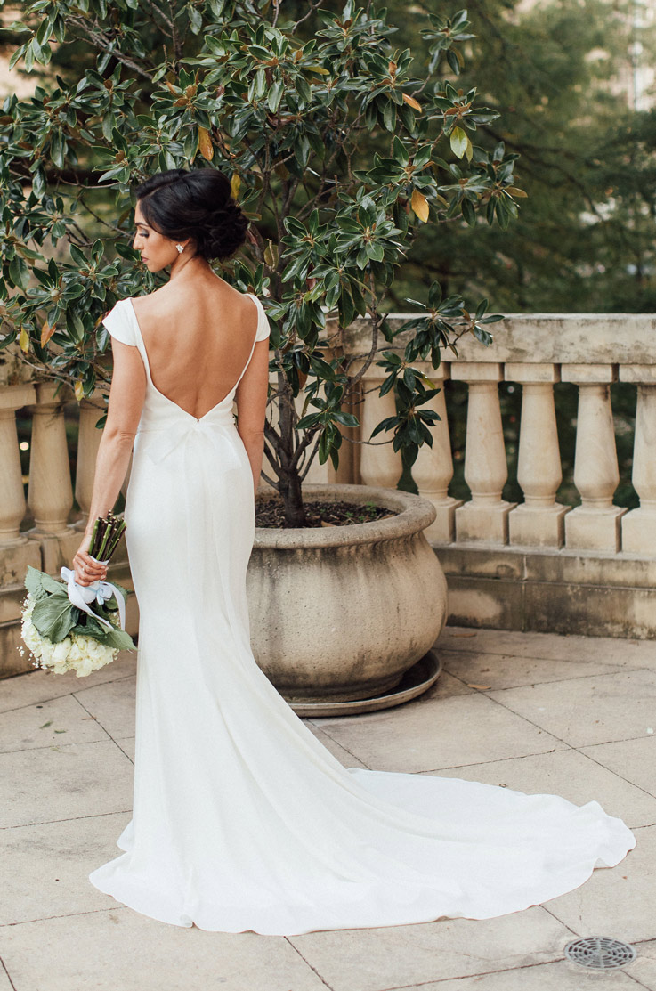Back of bride in a fit and flare crepe gown with a low back, stands on a terrace and holds a bouquet to the side.