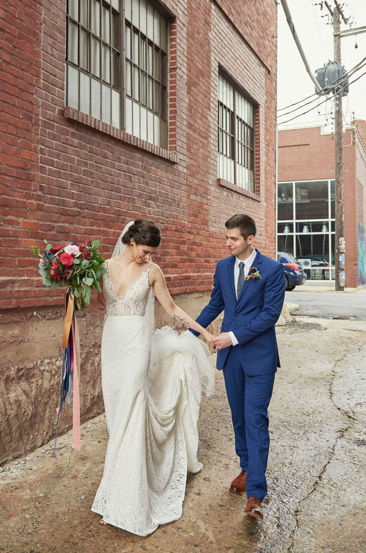 Bride in a V-neckline fit and flare wedding gown and veil, walks in a brick alley and holds a bouquet beside the groom.