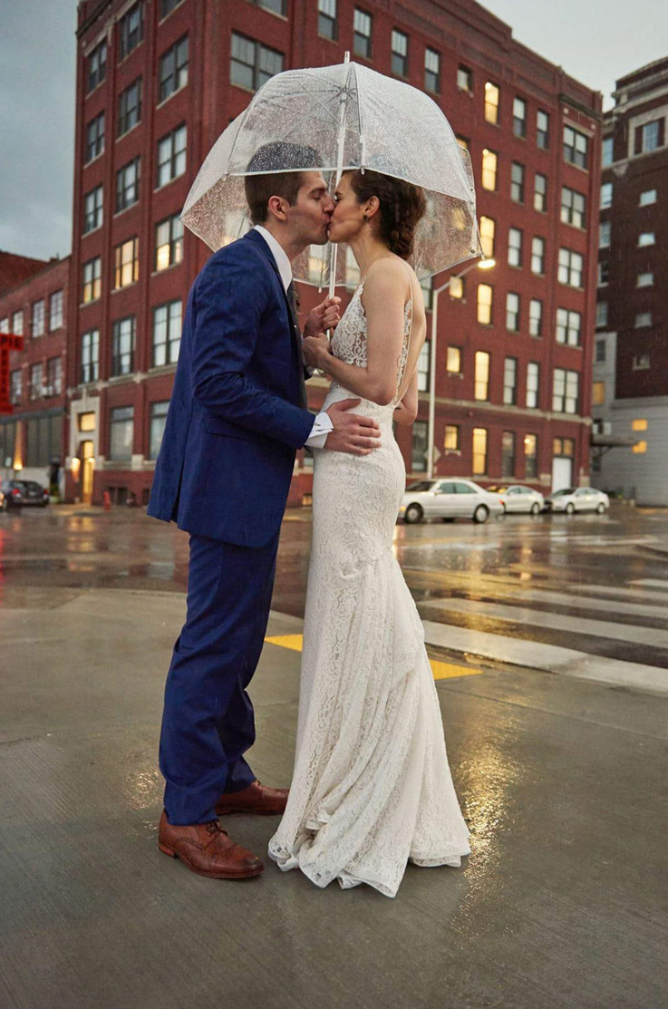  Bride in a fit and flare lace gown with a deep V-neckline, kisses groom in the rain under a clear umbrella on a city street.