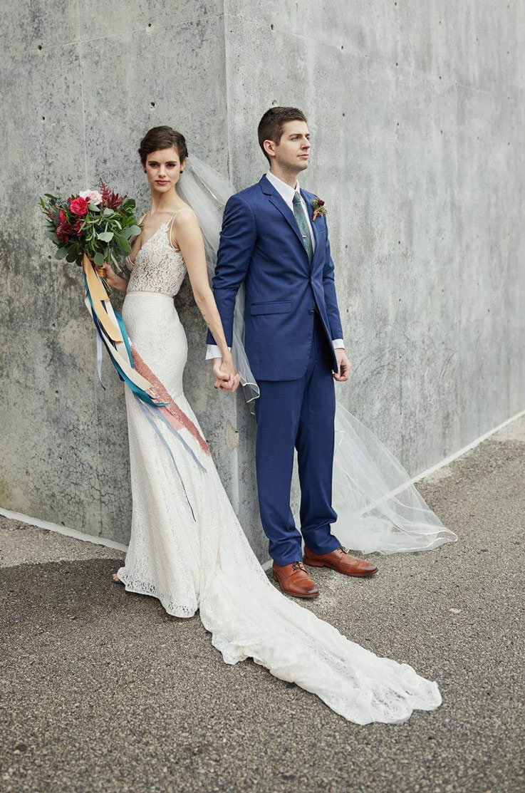 Bride in a fit and flare sleeveless gown poses and holds hands with the groom in a blue suit, against a concrete wall.
