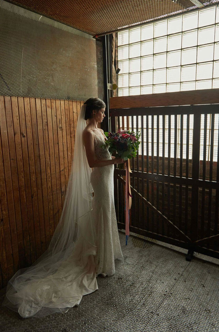 Bride in a fit and flare lace gown with a flowing veil, holds a bouquet and poses by an industrial window, in dramatic lighting.
