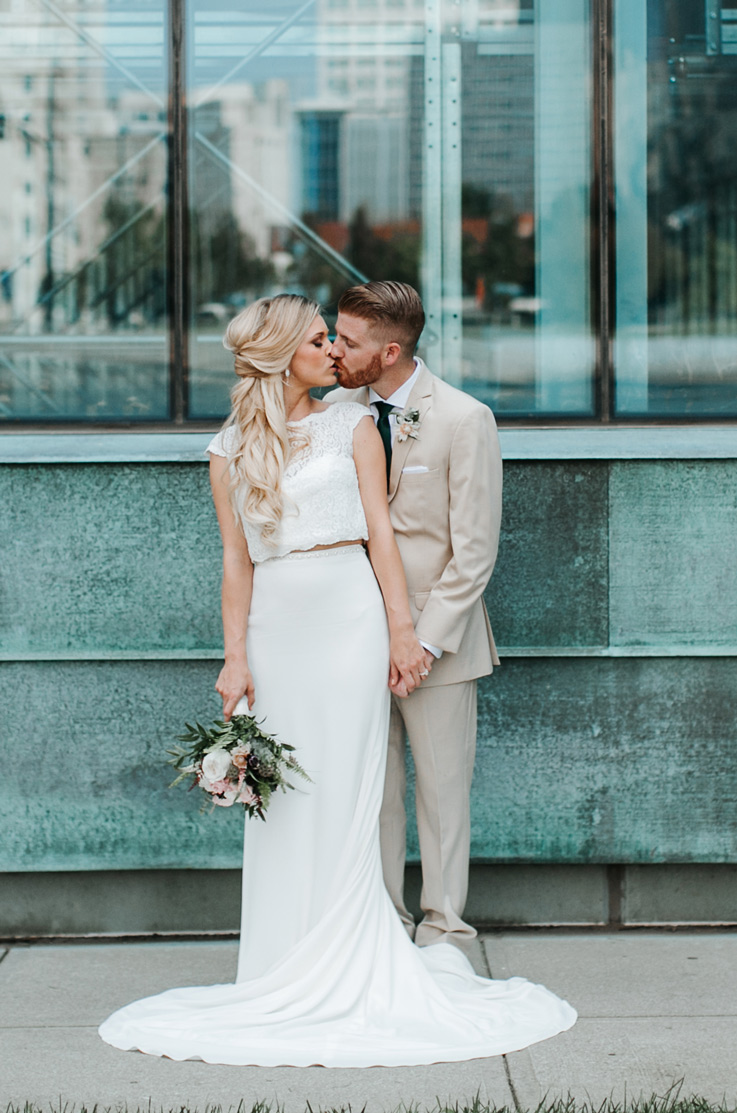 Bride in a two-piece wedding gown with a cropped lace top and fit and flare skirt, holds a bouquet and kisses groom.