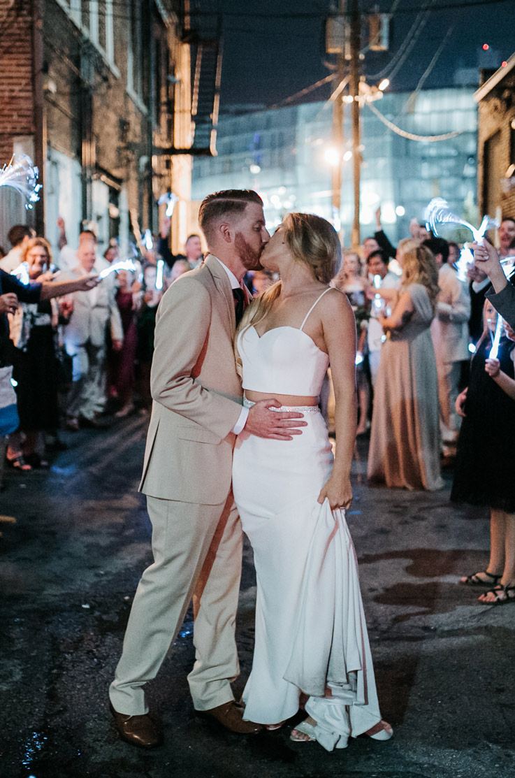Bride in a two-piece wedding dress with a fitted skirt and cropped bodice, kisses the groom with sparklers around them at night. 