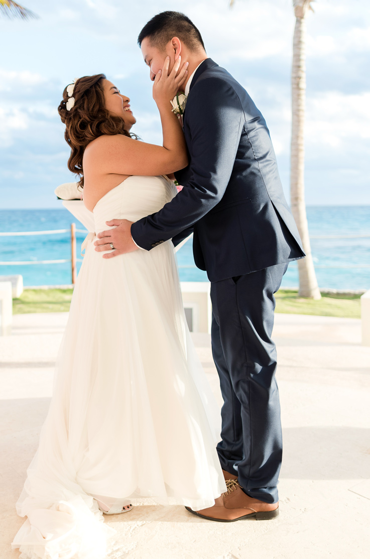  Bride in a strapless tulle wedding gown with a sweetheart neck, ribbon and flowing skirt faces the groom at a beach venue.