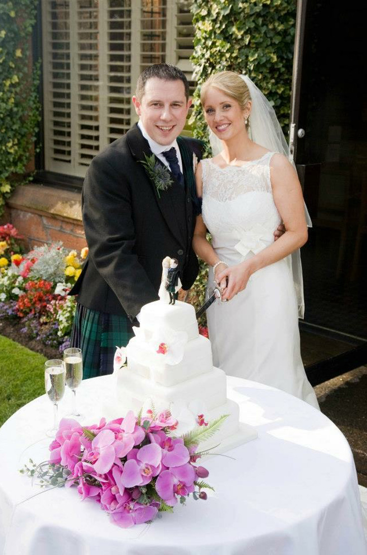 Bride in an elegant lace ivory gown, cutting cake with groom in garden setting, surrounded by bright flowers and outdoor decor.