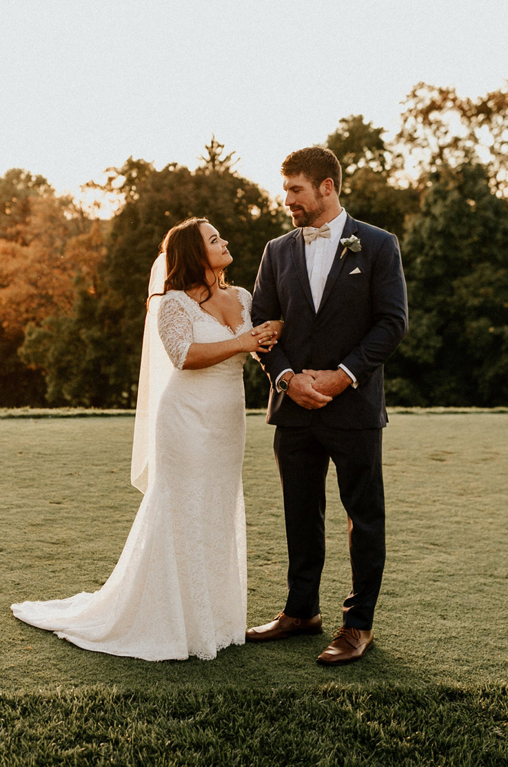 Bride in a lace fit and flare gown with a chapel train, a plunging neck and veil, links arms with and faces the groom.