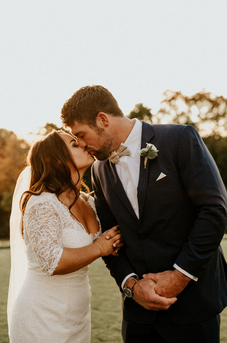 Bride in a lace sleeve gown with a plunging neckline and veil, kisses the groom in a dark suit, outdoors in the sunlight.