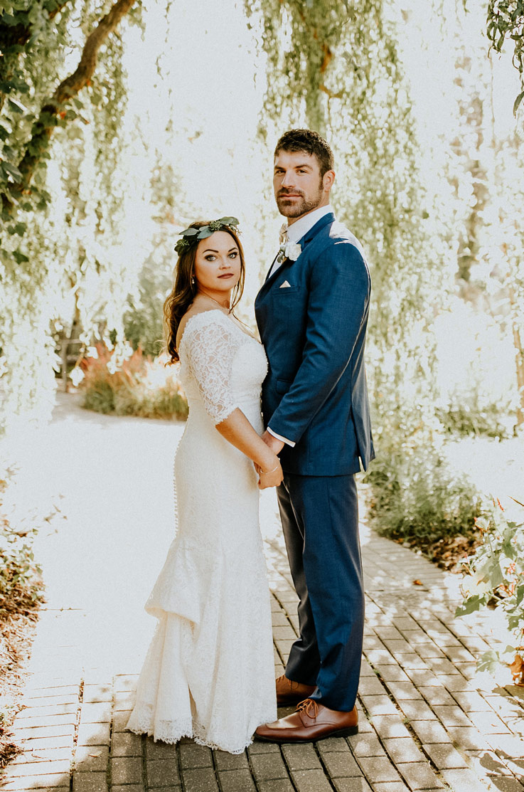 Bride in a lace fit and flare wedding dress with a low back, holds hands and poses with the groom under a willow tree.