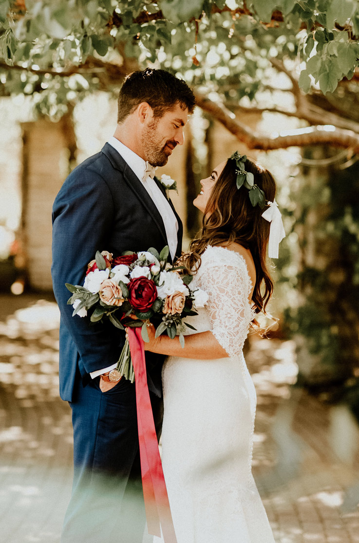 Bride in a fit and flare lace wedding dress with a plunging neck and sleeves, looks up at groom and holds a bouquet under a tree.