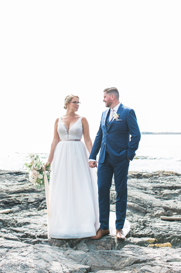 Bride in a lace bodice wedding dress with ribbon and a tulle skirt, stands on a rocky shoreline, holding hands with the groom.