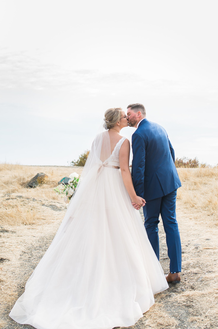 Bride in a sleeveless lace gown with a tulle skirt and open back with ribbon and a veil, kisses the groom on a coastal clifftop.
