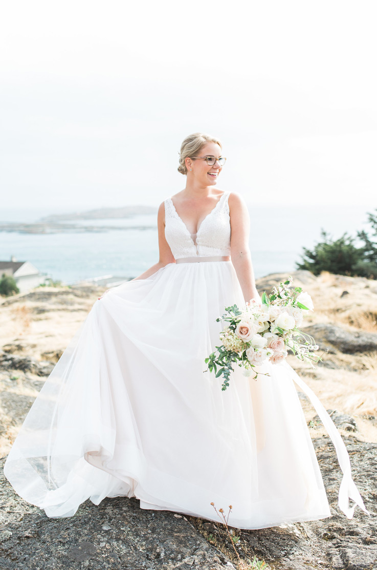 Smiling bride poses in a plunging neck gown with a tulle skirt, ribbon and lace bodice, holding a blush bouquet on a clifftop.