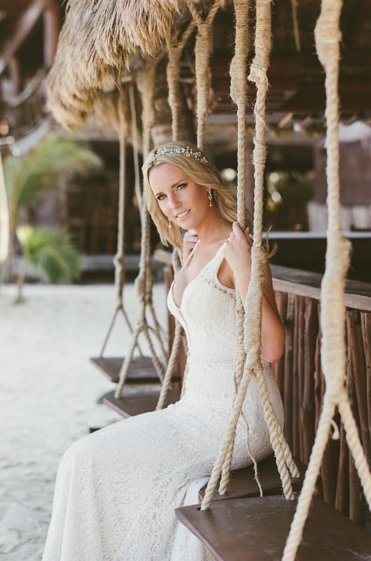  Bride in a lace fit and flare wedding dress with a plunging neckline and beaded waistband, seated on a swing at beachfront.