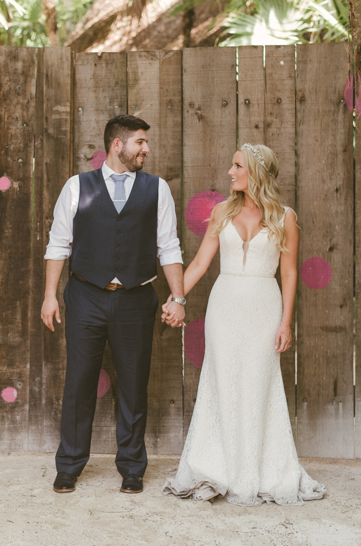 Bride in a lace fit and flare wedding gown with a beaded belt and plunging neck, stands outdoors and holds hands with groom. 