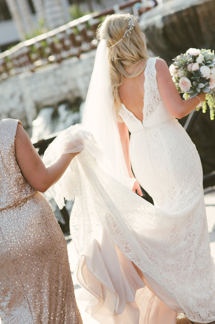 Bride in a deep-V back lace gown with a beaded belt and chapel train held by a bridesmaid, while holding a blush bouquet. 