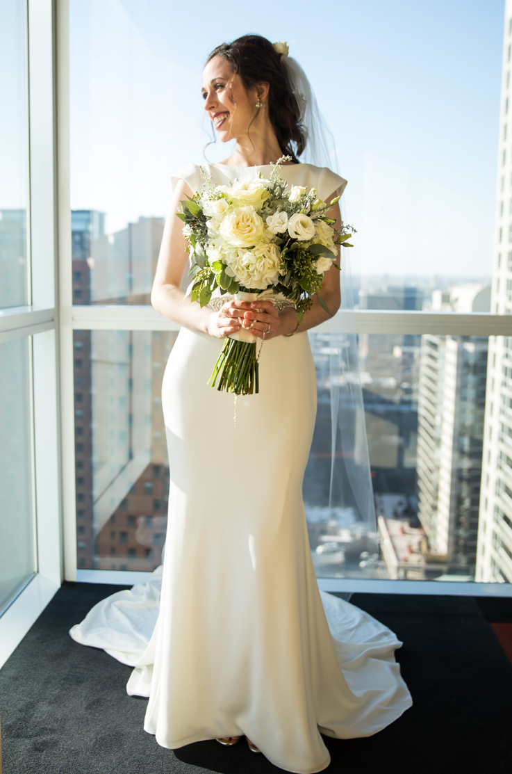 Bride in a crepe fit and flare wedding dress with cap sleeves and a veil, holds a bouquet indoors by tall glass windows.