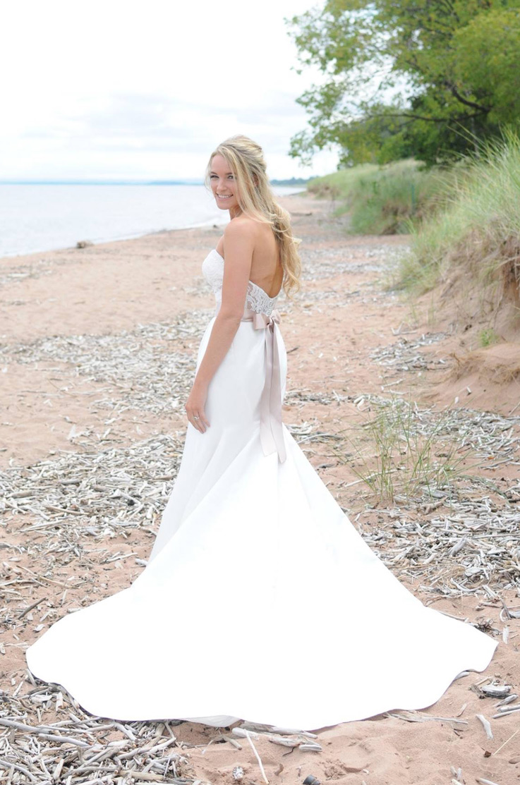 Bride in a strapless lace gown with ribbon at the waist, an open back and cathedral train, poses on a beachfront by the shore.