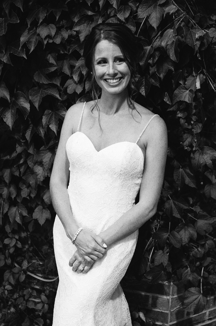  Smiling bride in a lace fit and flare gown with beaded spaghetti straps, against an ivy-covered wall in black and white.