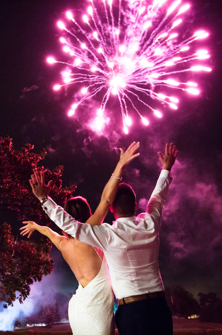 Bride in a backless lace fit and flare wedding dress, celebrating with groom outdoors at night under colorful fireworks.