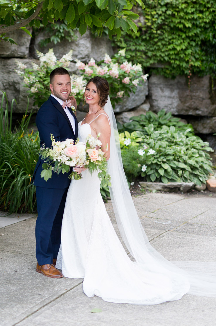 Bride in a fit and flare lace gown with a sweep train and veil, smiles next to her  groom with a light pink bouquet.