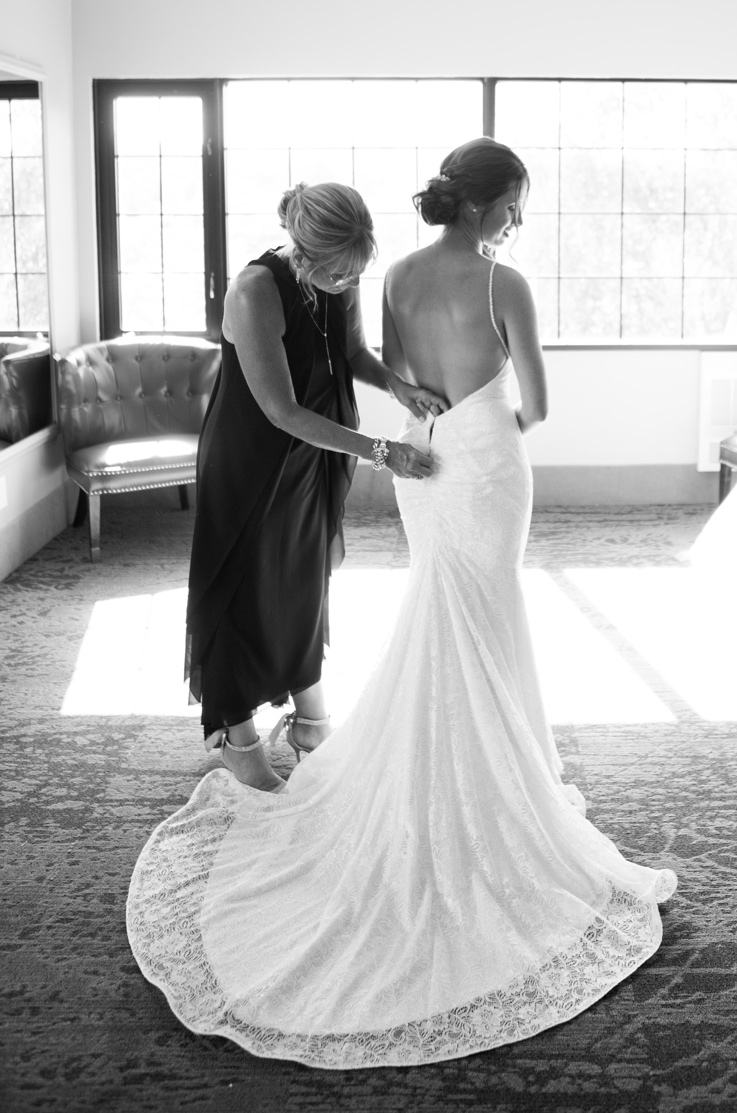 Back of bride being fastened into lace fit and flare wedding dress with a flowing sweep train, indoors by a bright window.
