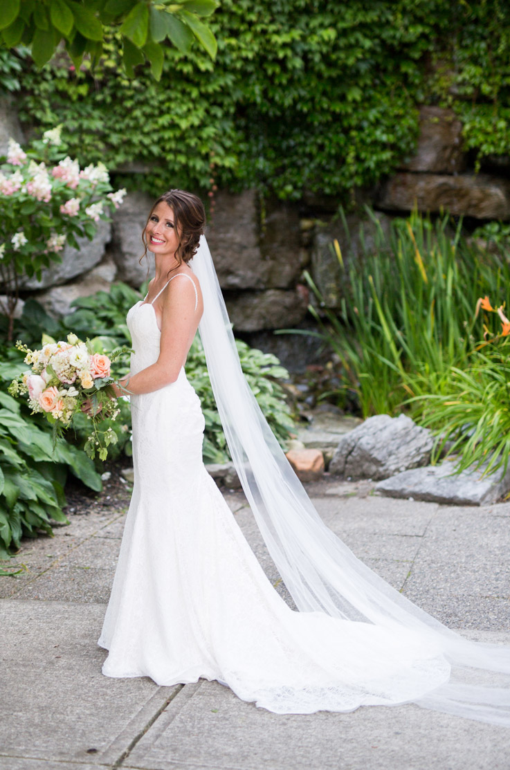 Bride in a fit and flare wedding gown with a sweep train and veil, is holding a bouquet and stands in a garden. 