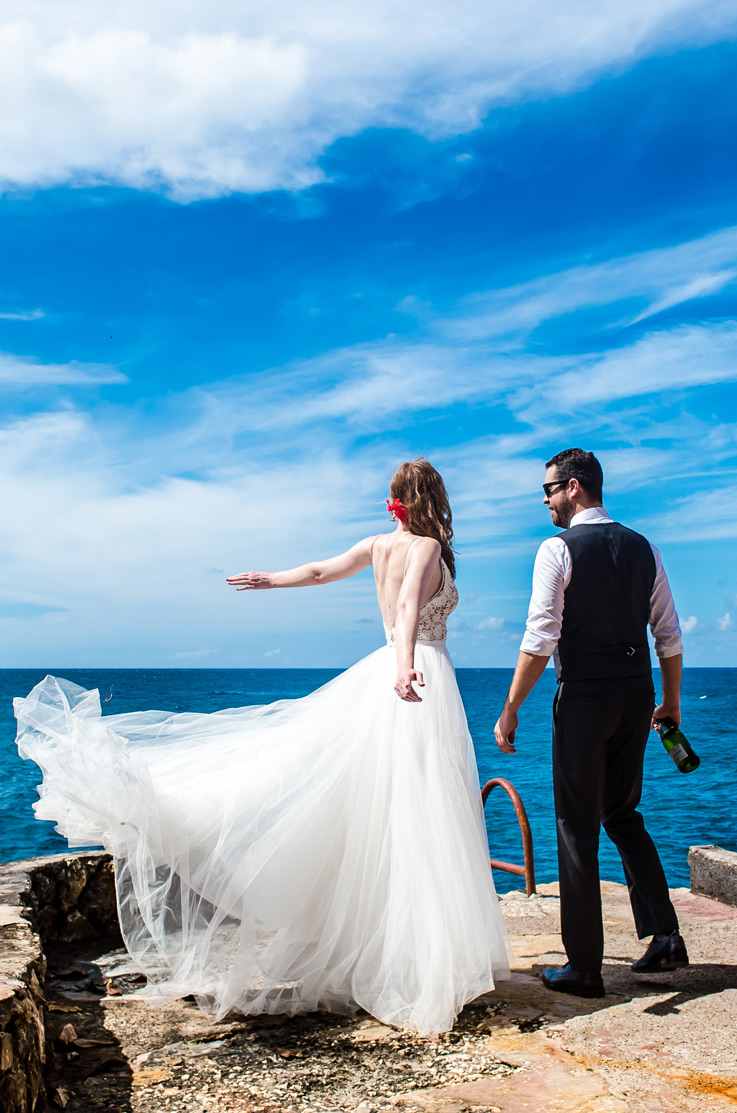 Bride in a flowing tulle wedding dress with a V-neck lace bodice, stands on a seaside cliff in ocean breeze next to groom.