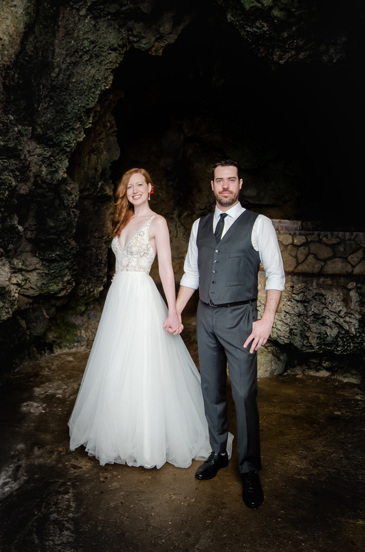 Bride in a lace bodice wedding gown lined in tulle, poses with groom and they hold hands in a rocky cave. 