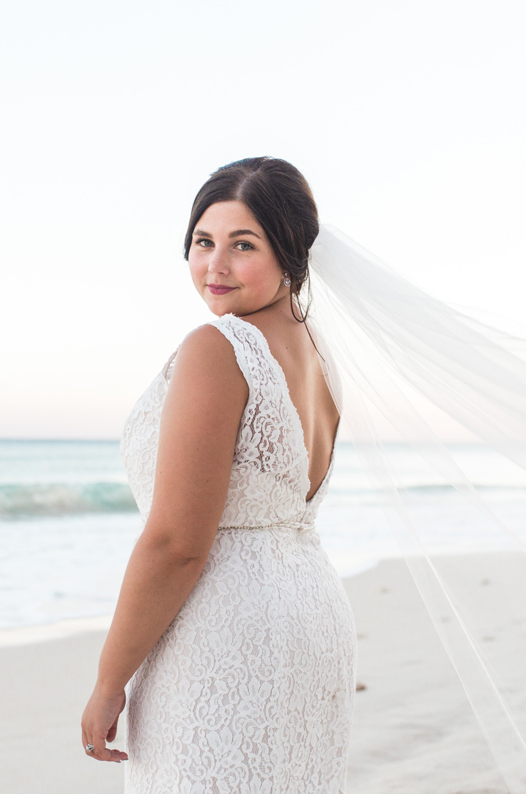 Bride in a sleeveless fit and flare gown in lace, with a beaded belt, open back and veil, poses on a beachfront.