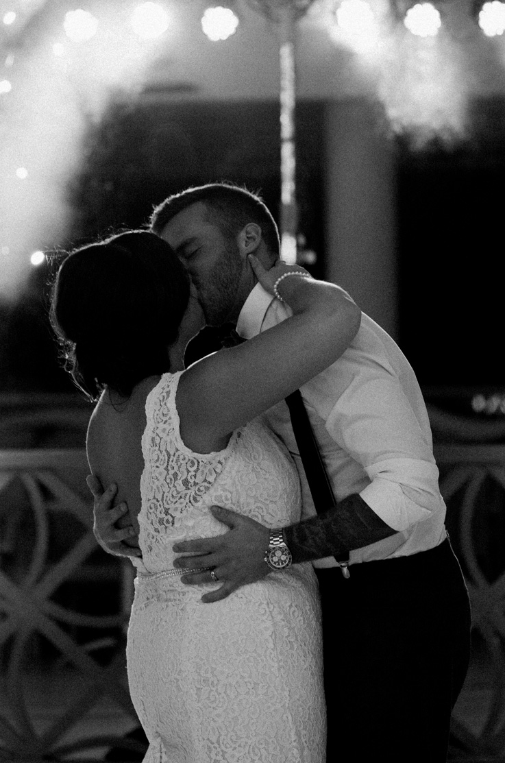 Bride in a lace gown with an open back and beaded belt, kisses groom on dance floor at night, in black and white.