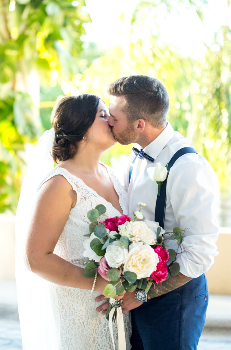 Bride in a lace plunging neck wedding gown with beaded belt detail and a veil, kisses groom outdoors and holds a bouquet.