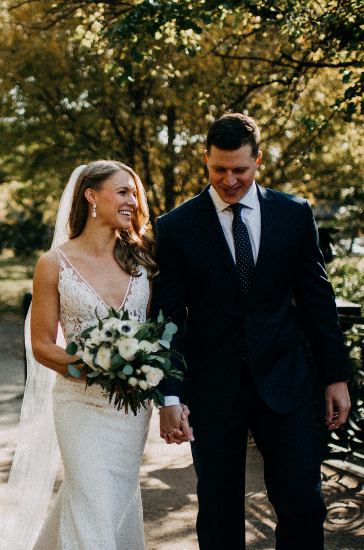 Bride in a lace V-neck gown with a veil, smiles and holds a bouquet with the groom as they walk outdoors, next to trees.