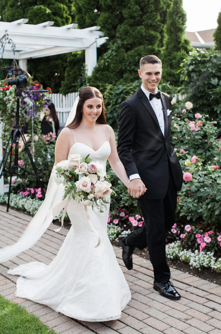 Bride in a fit and flare lace gown with a sweep train and veil, walks hand in hand with groom and a bouquet on a garden path.