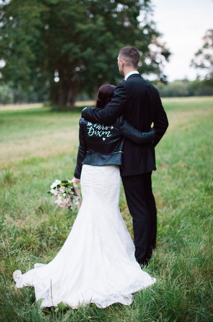 Bride in a lace fit and flare gown with a leather jacket, stands in a grass field with the groom and is holding a bouquet.