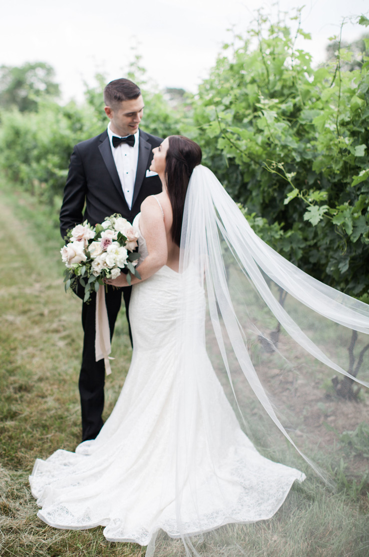 Bride in a low back fit and flare gown with a veil and sweep train, looks up at the groom and holds a bouquet in a vineyard
