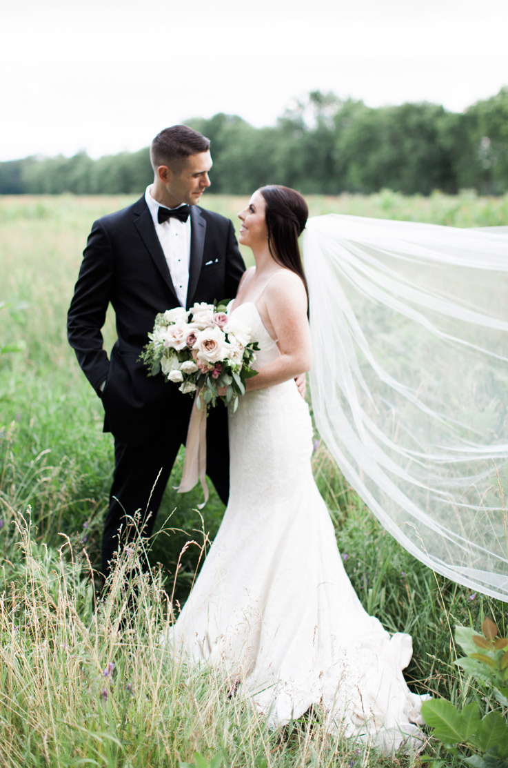 Bride in a fit and flare gown with a veil and thin straps, stands in a grassy field with the groom and holds a bouquet.