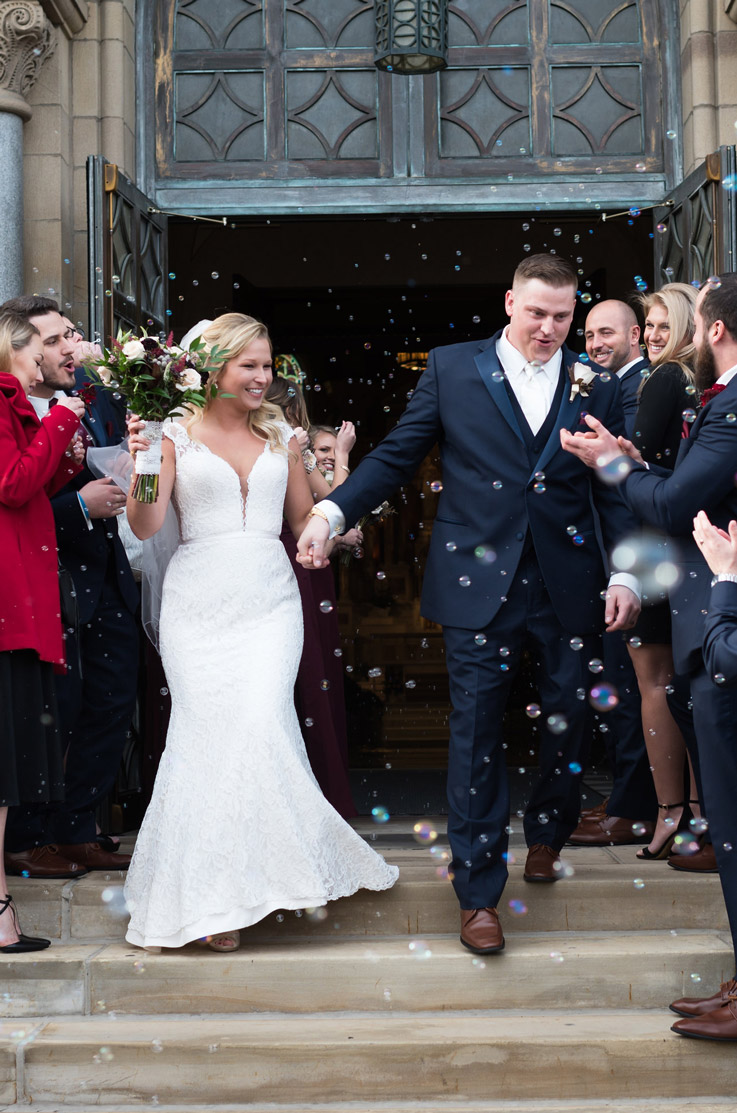 Bride in a fit and flare lace gown with cap sleeves, exits the venue with bubbles and holds hands with the groom.