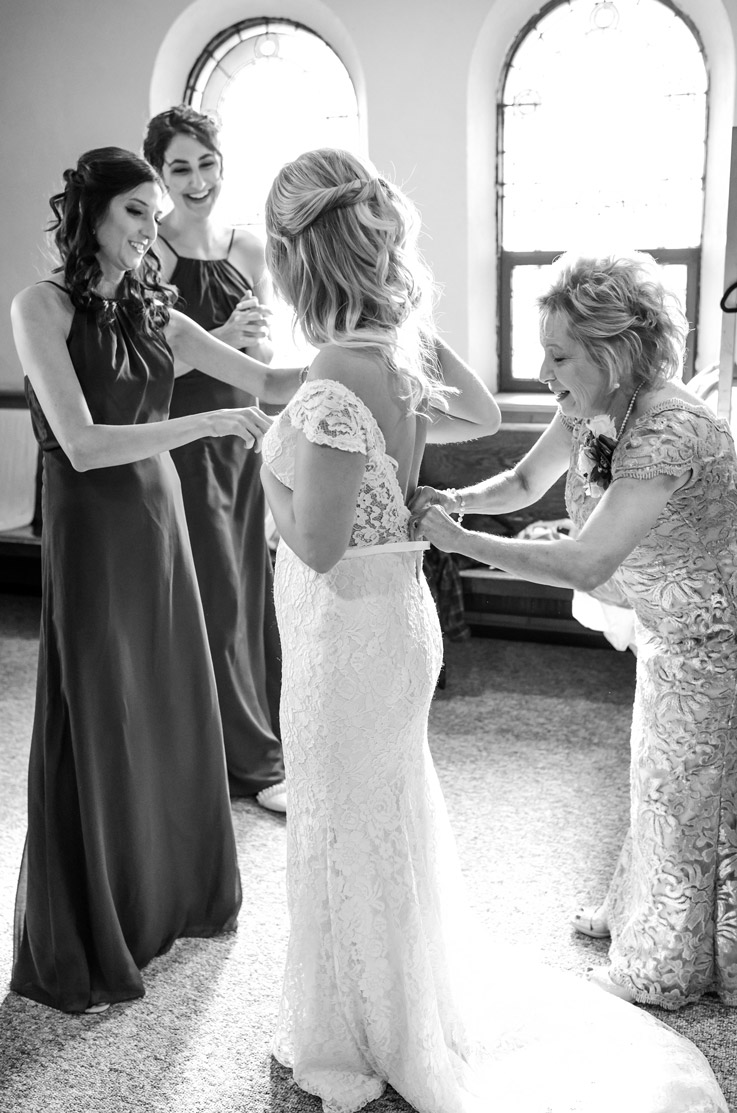 ride being fastened into a lace low back gown with cap sleeves, and is surrounded by bridesmaids indoors, in black and white. 