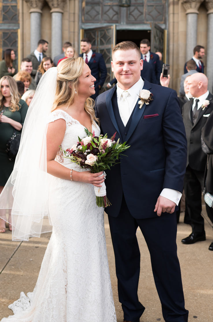 Bride in a plunging neck lace gown with a veil, holds a bouquet and smiles at the groom in front of the venue and guests.