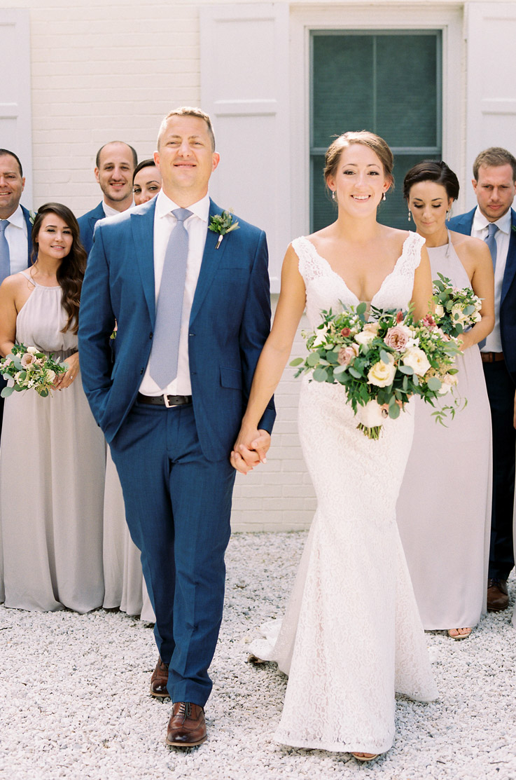 Bride in a lace fit and flare wedding dress with a plunging neck, walks hand in hand with groom past guests and with a bouquet.