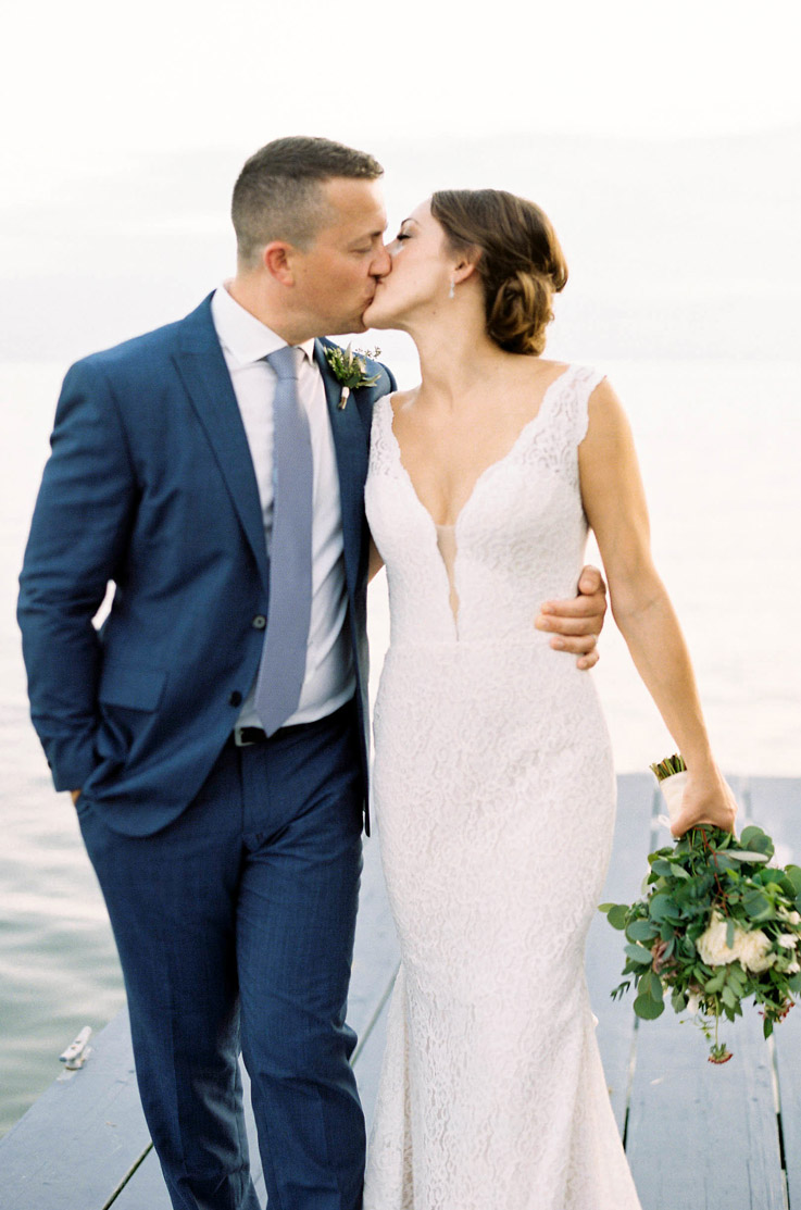 Bride in a fit and flare lace gown with a plunging neckline, kisses the groom in a blue suit and holds a bouquet by the ocean.