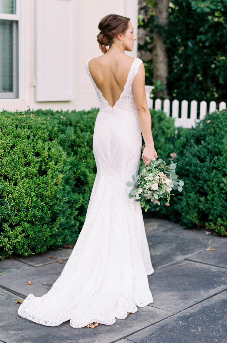 Back of bride in a low back fit and flare gown with a chapel train, holding a bouquet and poses on a garden patio.