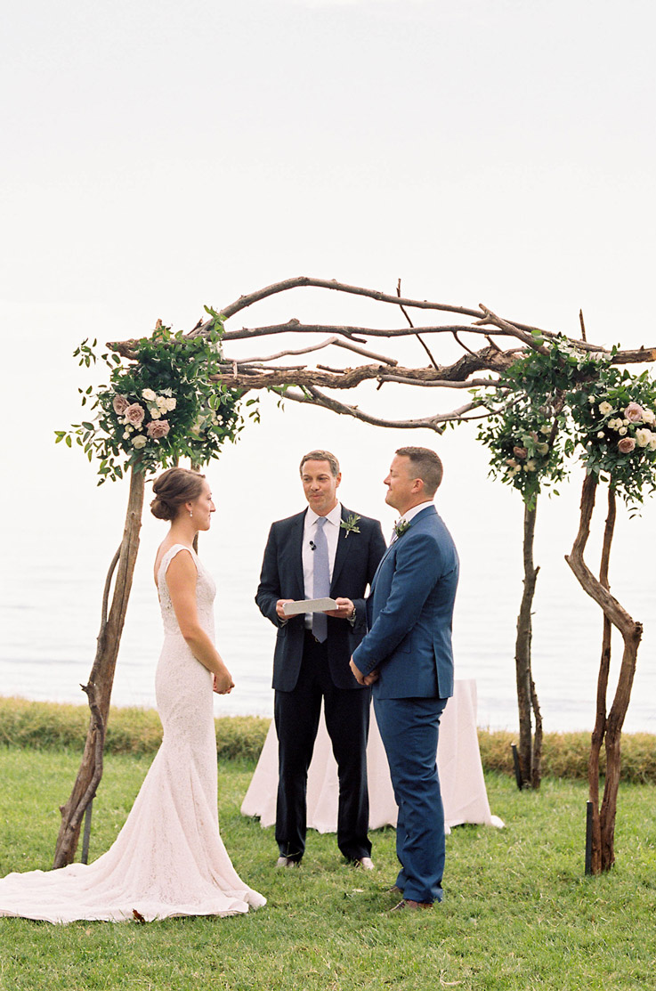 Bride in a lace fit and flare gown with a flowing train, faces the groom in a blue suit at the altar, on a waterfront.