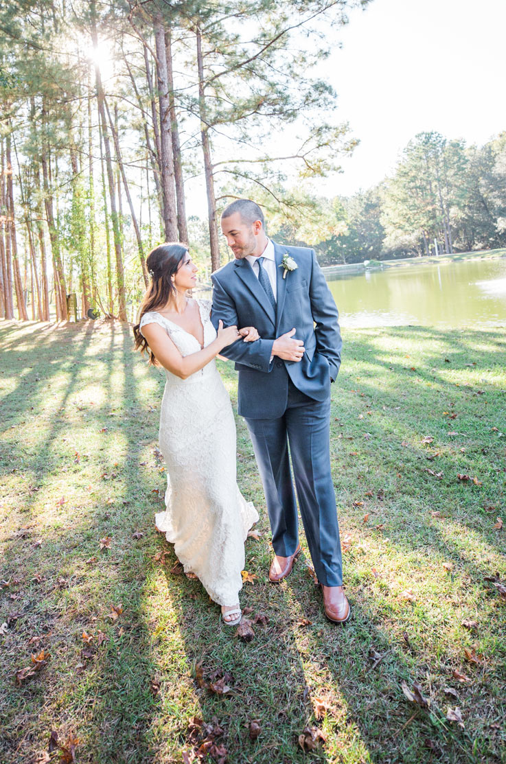 Bride in a plunging neck lace wedding dress with cap sleeves, walks with groom outdoors in a sunlit area with tall trees.