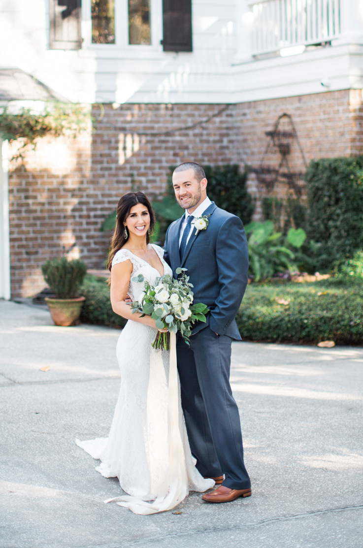Bride in a fit and flare lace gown with cap sleeves, poses with the groom outside the venue with a bouquet. 