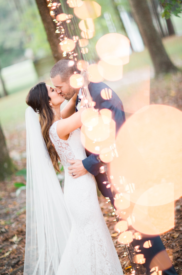 Bride in a lace fitted wedding dress with cap sleeves and a veil, kisses groom in a blue suit in a forest with lights.