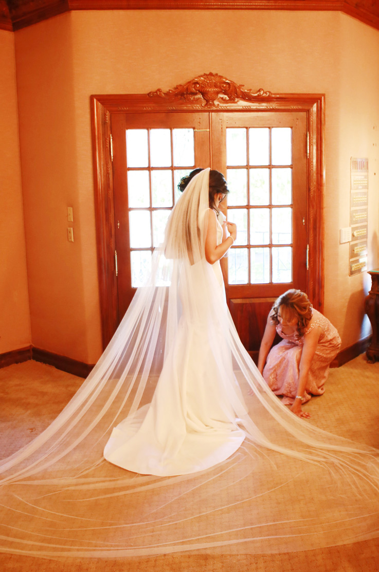 Bride in a fit and flare satin gown with a chapel train and veil, gets ready with a bridesmaid by the entrance. 