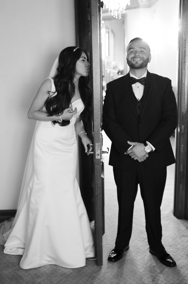 Bride in a satin fit and flare gown opens a door, next to the groom on the other side of it posing in a black tuxedo.