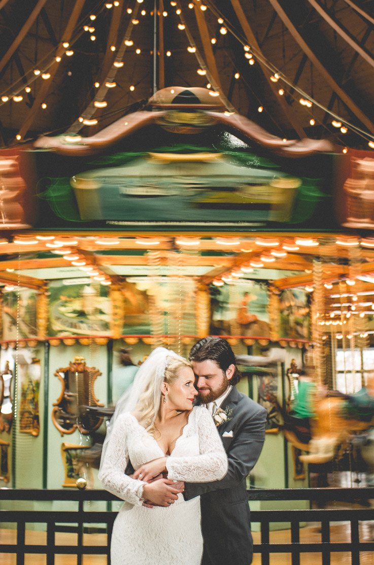 Groom embraces bride wearing a plunging neck lace gown with long sleeves and a veil, in front of a carousel with lights. 